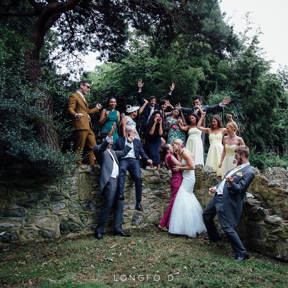 Wedding group portrait, couple kissing, surrounded by jubilant guests on stone steps in lush greenery.