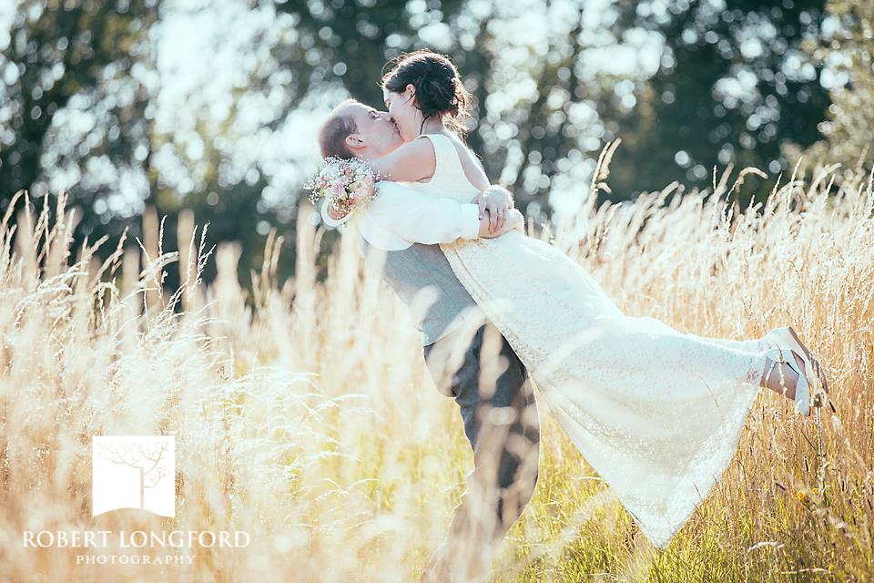 Newlyweds embrace and kiss in a field of tall grass. The groom lifts the bride.