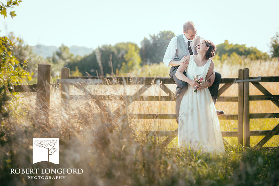 Bride and groom playfully pose on a wooden fence in a sunny field.