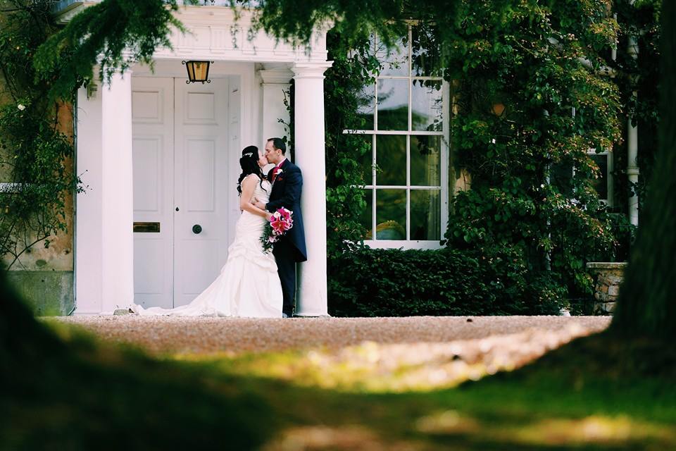 Couple embraces in front of a white building with columns; woman in a wedding dress, man in a suit, both looking at each other.