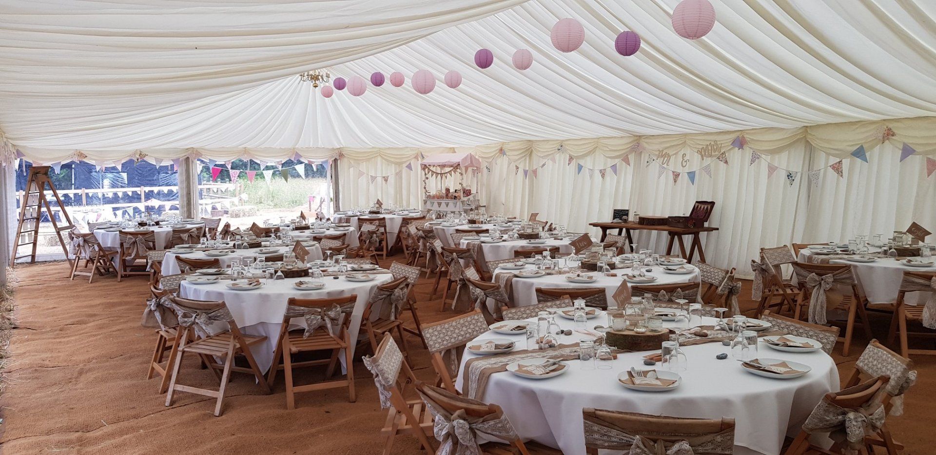Inside a decorated tent, tables are set for a gathering; pink paper lanterns hang overhead.