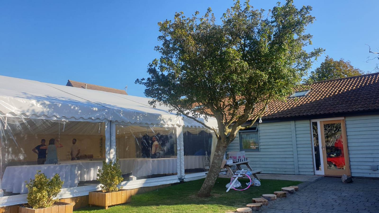Event venue with a white tent, tree, and wooden building. Sunny day.