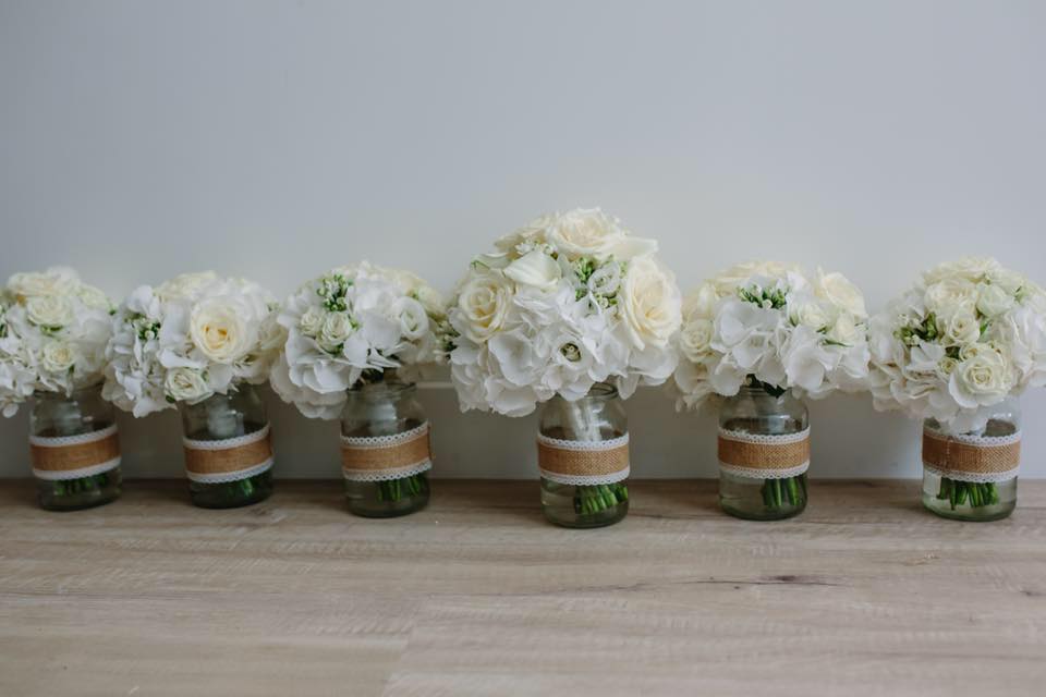 Six white floral bouquets in mason jars wrapped with burlap ribbon.
