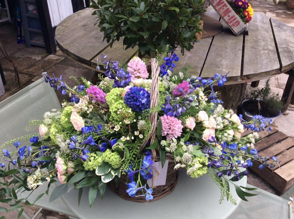 Basket of colorful flowers on a table. Includes blue, pink, green, and white blooms.
