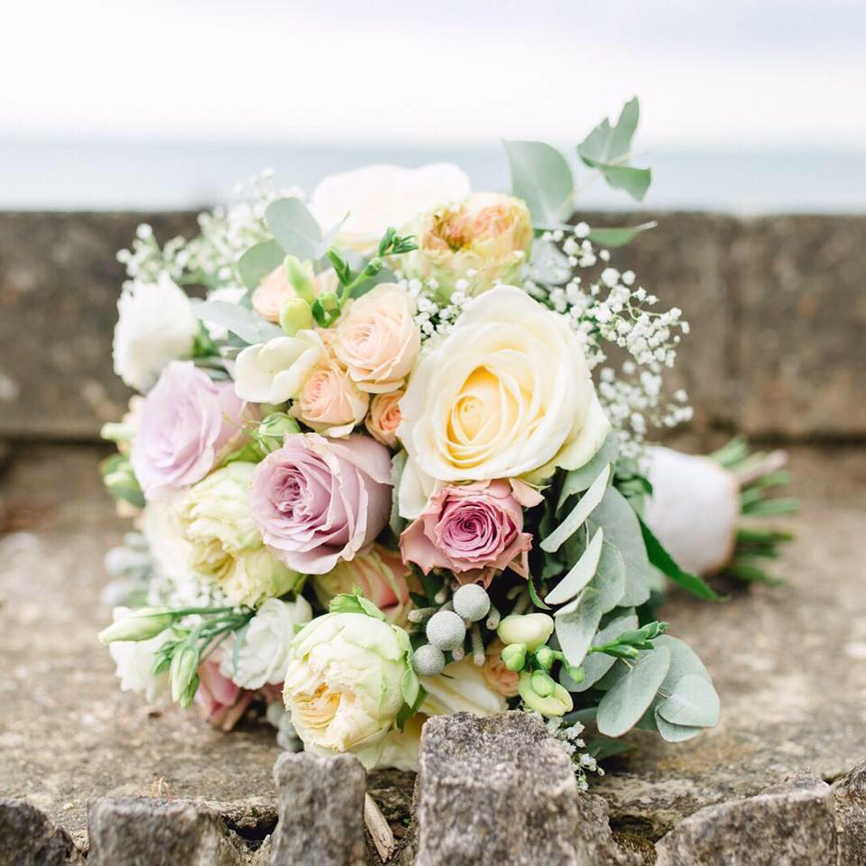 Bouquet of pastel roses and greenery on a stone wall, ocean in the background.