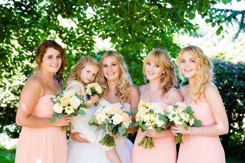 Bride and bridesmaids in peach dresses holding bouquets, smiling, posing under green trees.
