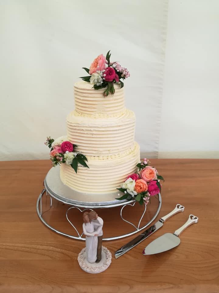 Three-tiered wedding cake with floral decorations, on a tiered silver stand. Couple figurine and cake-cutting set next to it.