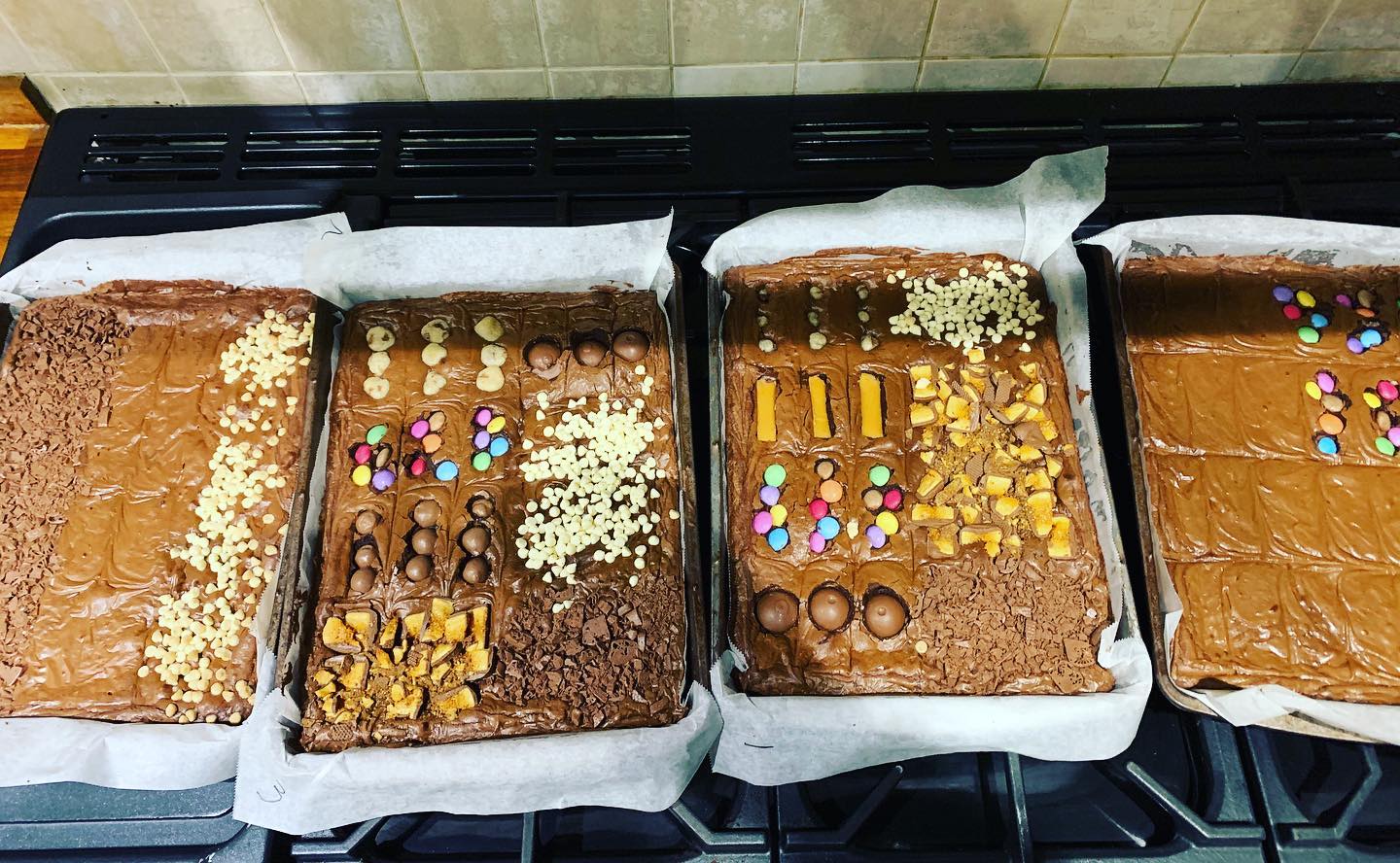 Four rectangular trays of brownies, each with different toppings, sitting on a stove.