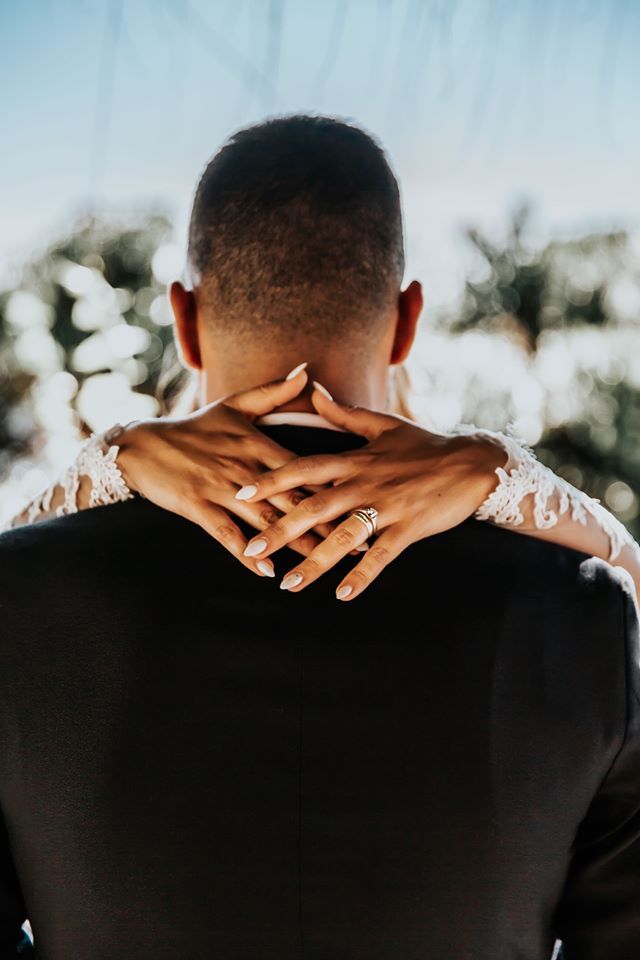 Bride's hands with lace sleeves clasped around groom's neck, outdoors.