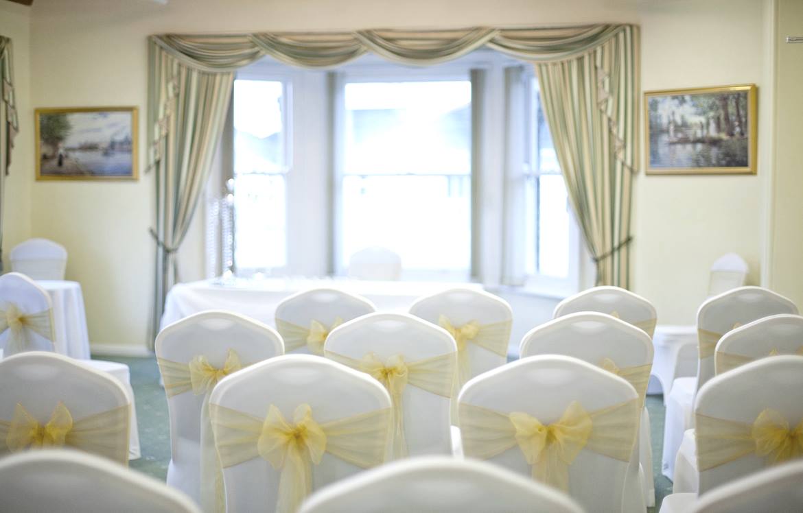 Wedding ceremony setup; rows of white chairs with yellow bows face a window, artwork on walls.