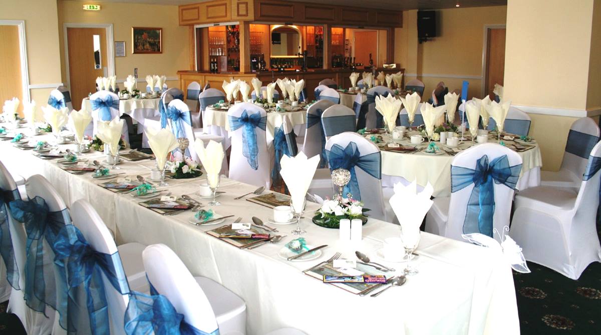 Banquet room with long tables set for a celebration, white linens, blue accents, and floral centerpieces.