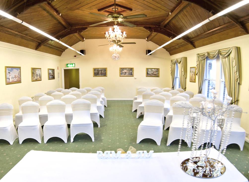 Wedding ceremony setup: white chairs, adorned, facing an altar in a room with a green carpet, and chandelier.
