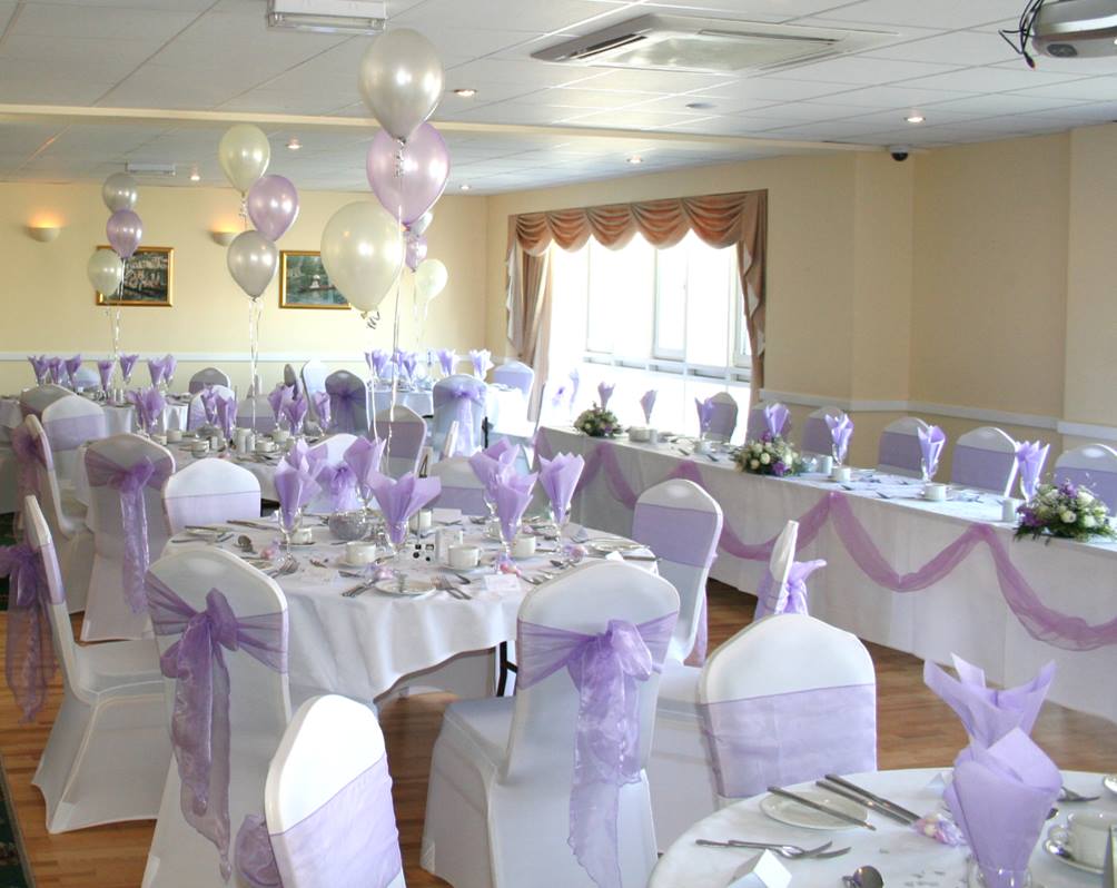 Wedding reception room decorated in white and lavender. Balloons, tables with centerpieces, and chair sashes.