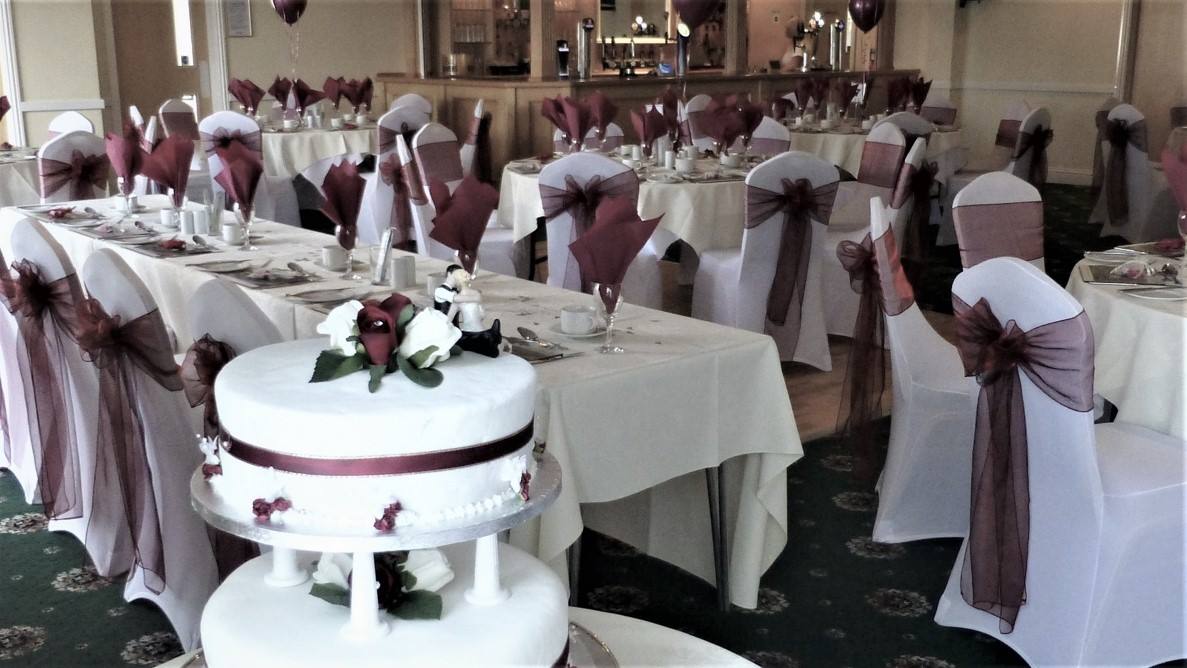 Wedding reception with tables set for guests, white tablecloths, and maroon chair sashes. A cake is in the foreground.