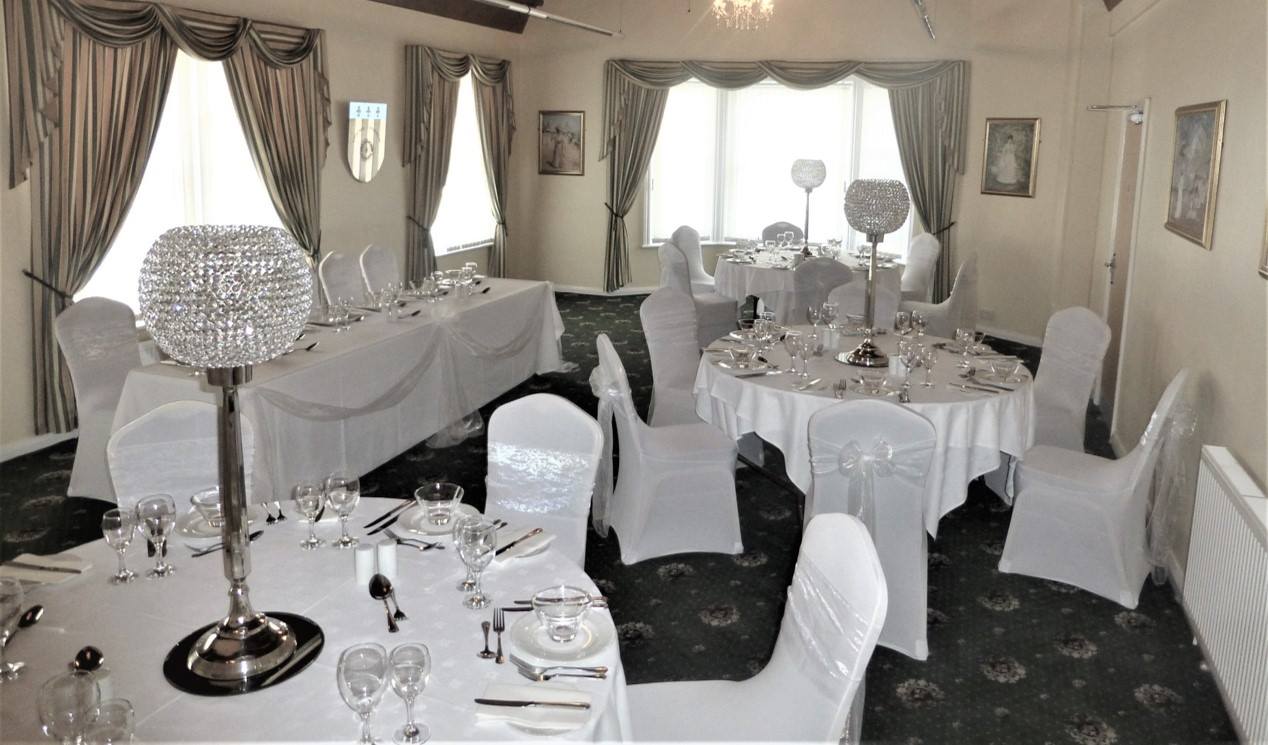 Formal dining room set for a celebration. Tables draped in white linens, adorned with centerpieces, chairs covered in white.