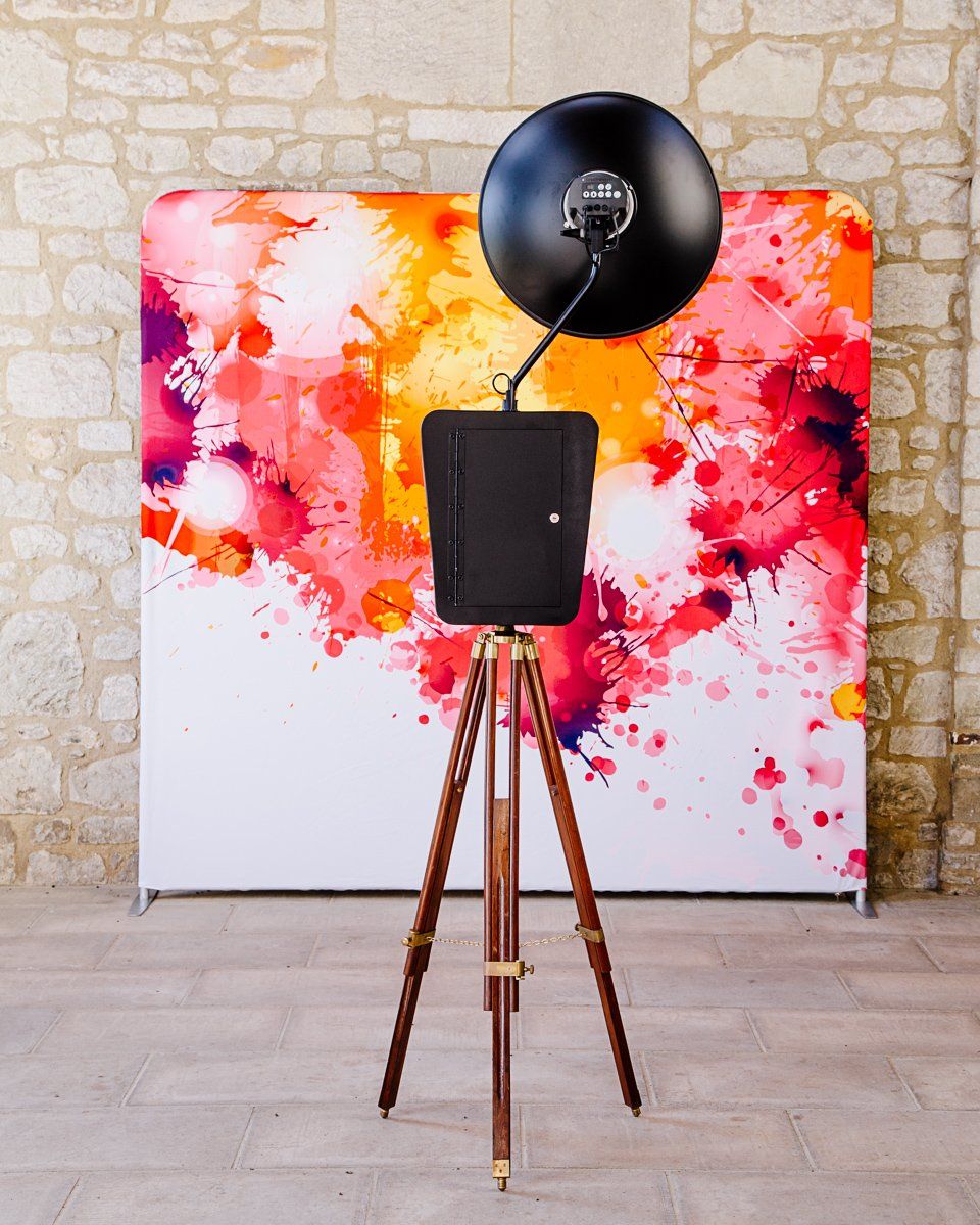 Photo booth with tripod and black camera, colorful splash art backdrop, set against a stone wall.