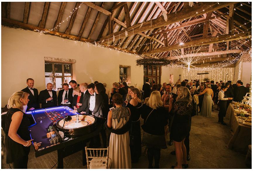 Wedding reception in a barn with guests in formal attire, fairy lights, and a food table.