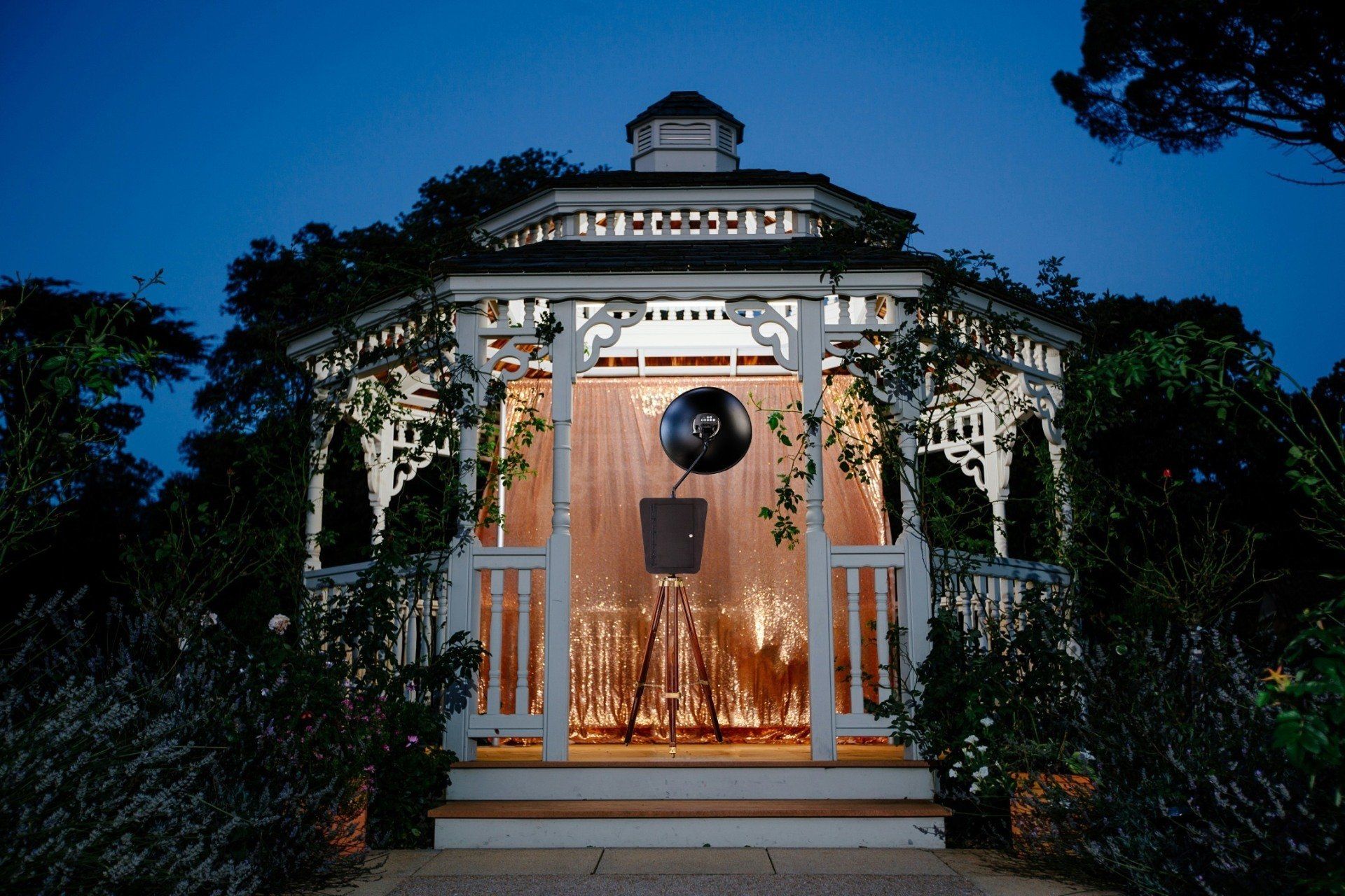 Lit gazebo at dusk, vines, fairy lights, table set.