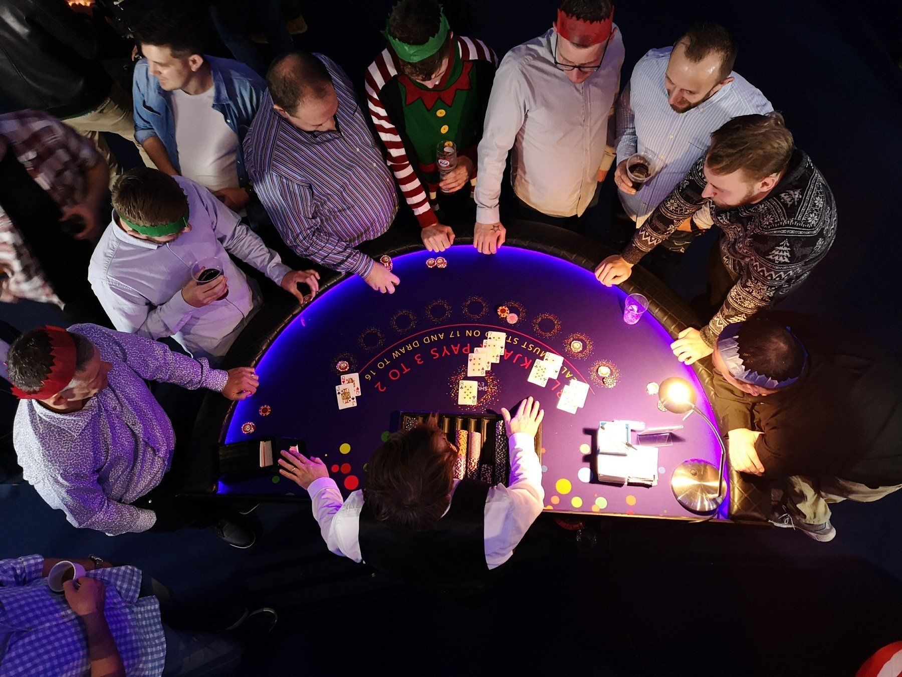 People around a poker table, illuminated, with cards and chips. Festive headbands, drinks.