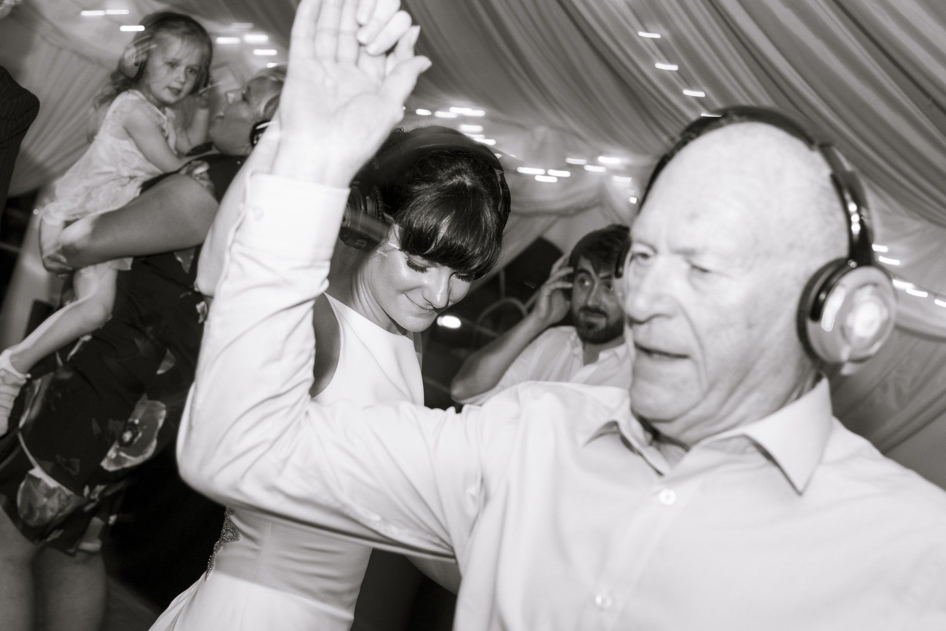 Man with headphones dances at a party; woman and child are nearby. Others look on, some smiling.