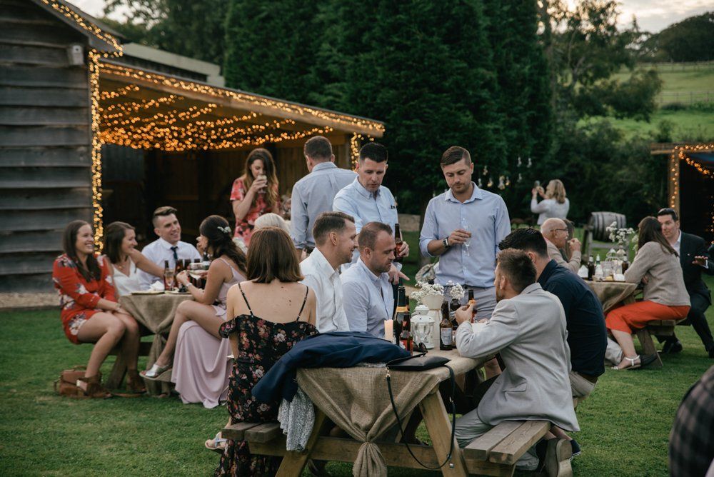 People seated at picnic tables outdoors, illuminated by string lights, likely at a gathering.