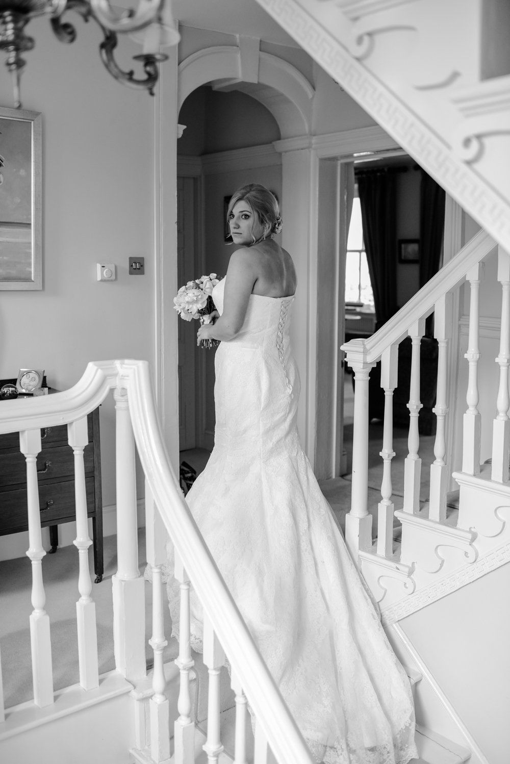 Bride in a white gown holding flowers, descending a staircase in a well-lit interior.