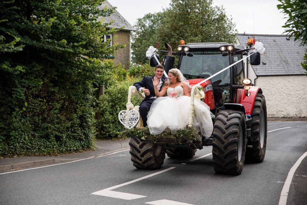 Newlyweds ride on a tractor decorated with flowers. The tractor is on a road, trees and buildings in the background.