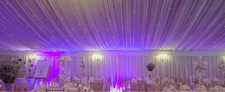 Elegant event space decorated with white draping, purple lighting, and tables set for guests.