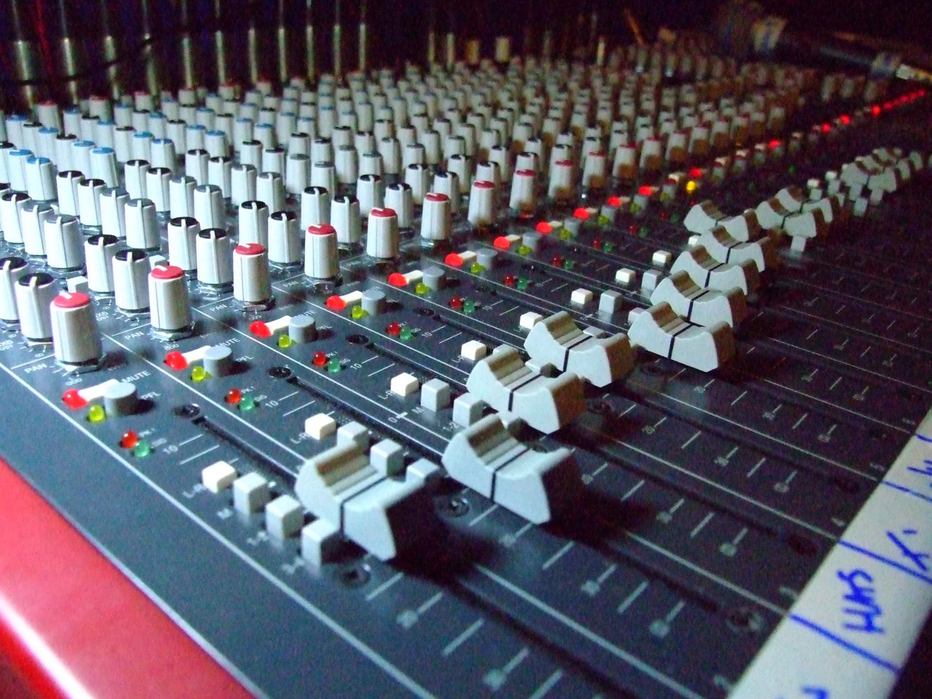 Close-up of a sound mixing console with multiple faders, knobs, and lights on a red surface.