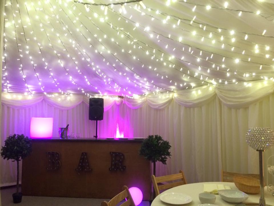 Bar area in a venue, decorated with fairy lights and purple uplighting. Includes a bar, speaker, and tables.