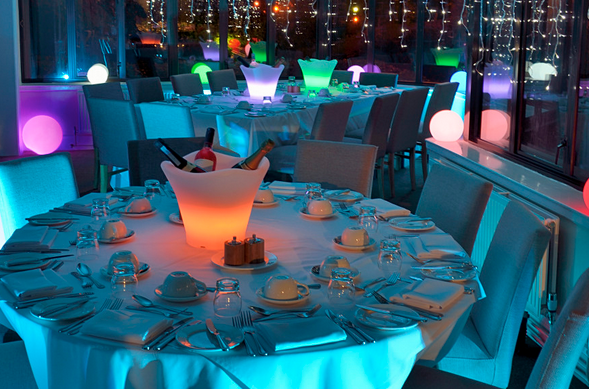 Round tables set for dining in a room with colorful mood lighting and illuminated ice buckets.