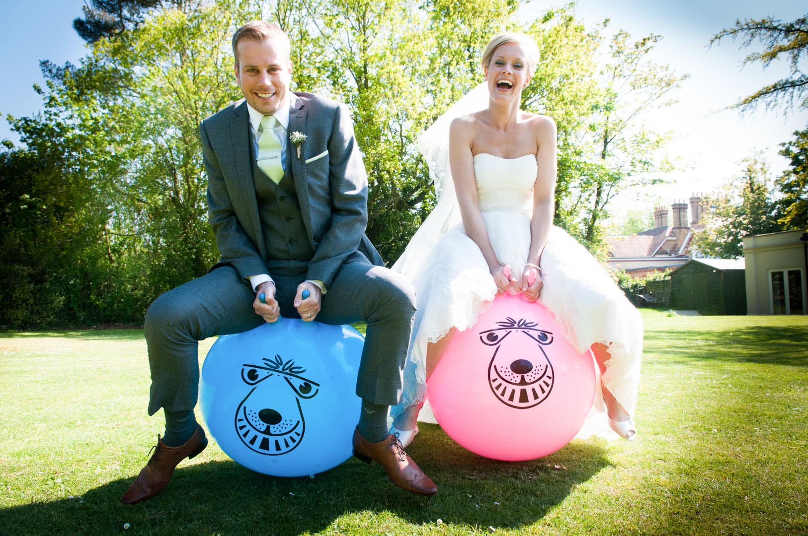 Bride and groom on space hoppers outdoors, laughing. Groom in gray suit, bride in white dress. Blue and pink balls.