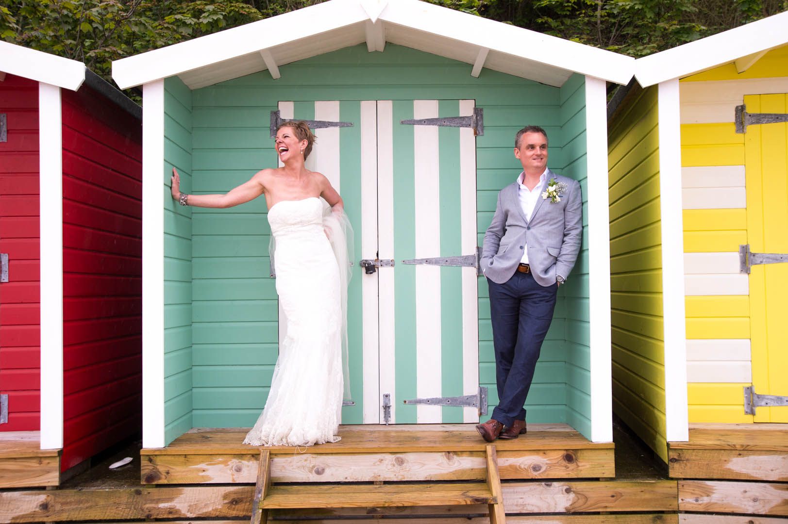 Newlyweds pose at a beach, standing in a mint-green and white striped beach hut; bride laughs, groom smiles.