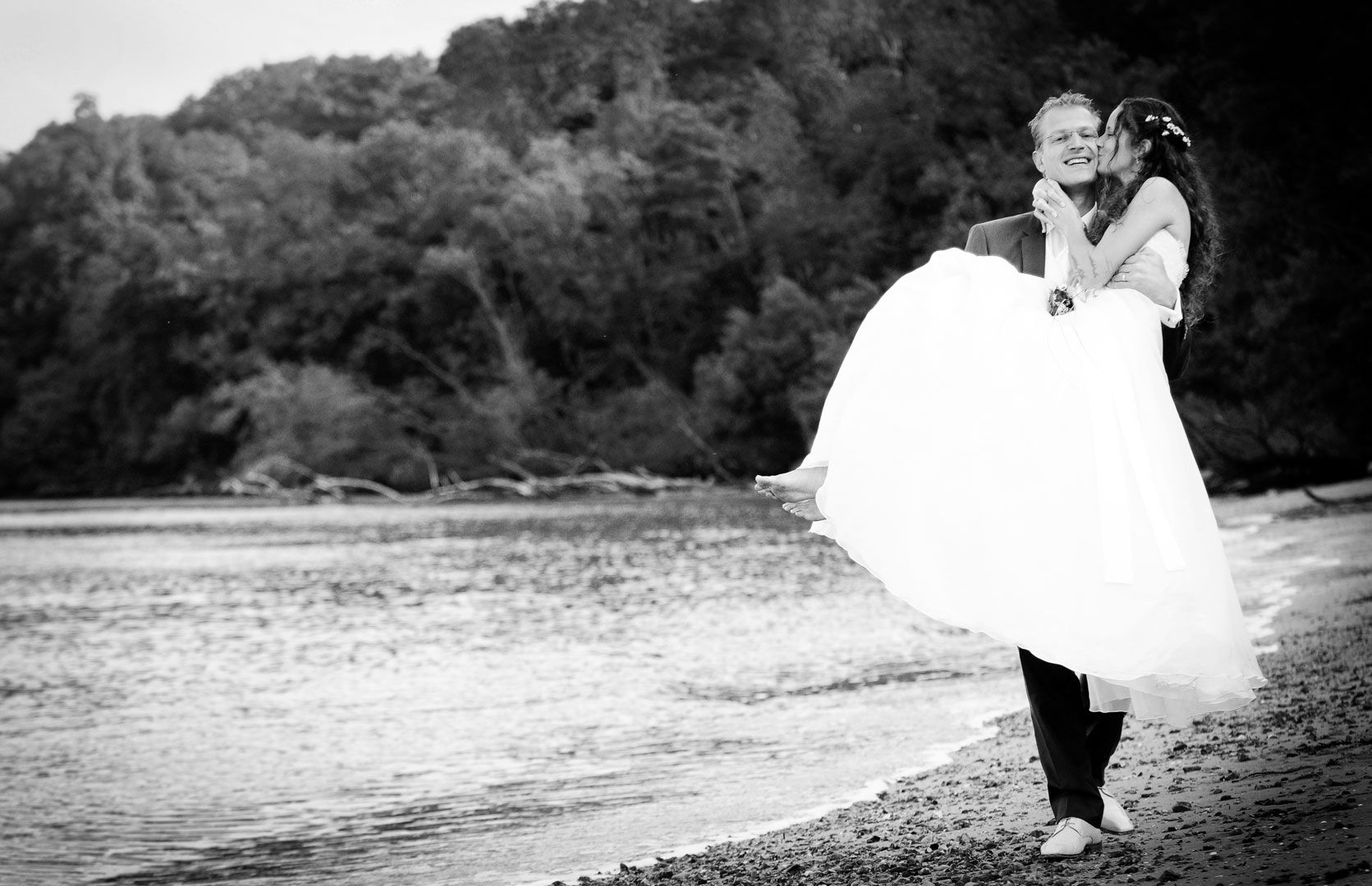 Groom carrying bride on a beach; she embraces him, smiling. Black and white photo with a forested backdrop.