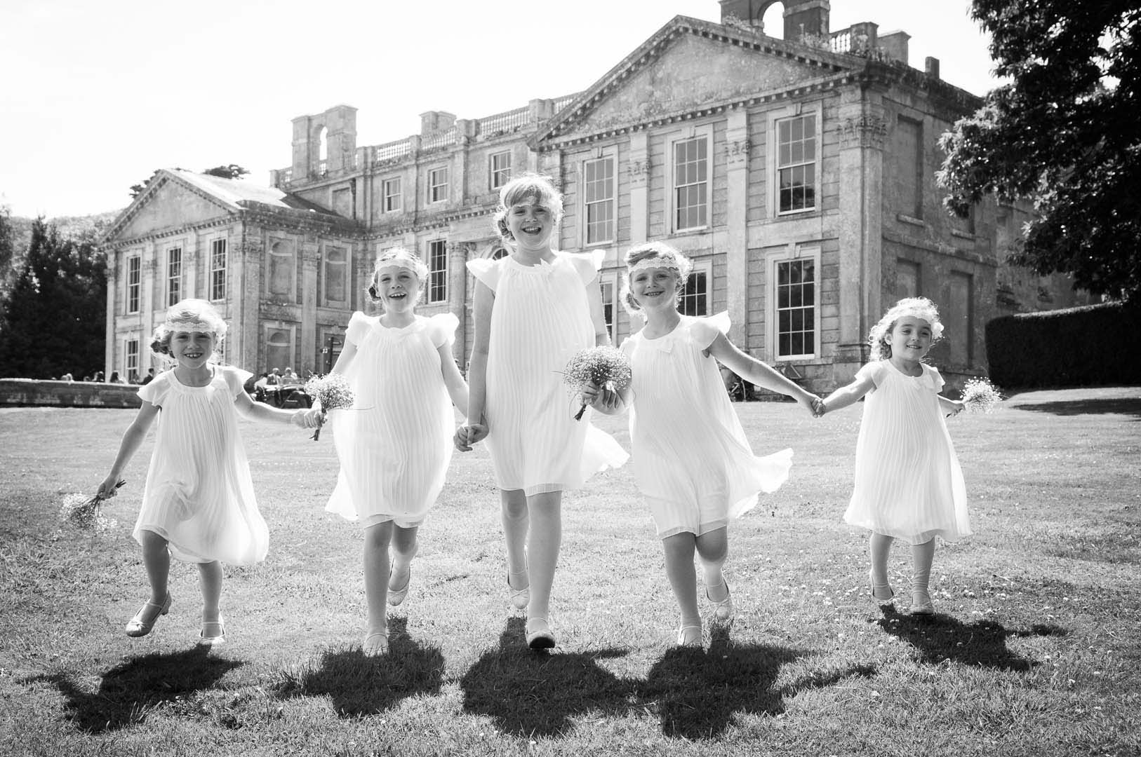 Five children in white dresses hold hands, running on grass in front of a large stately building.