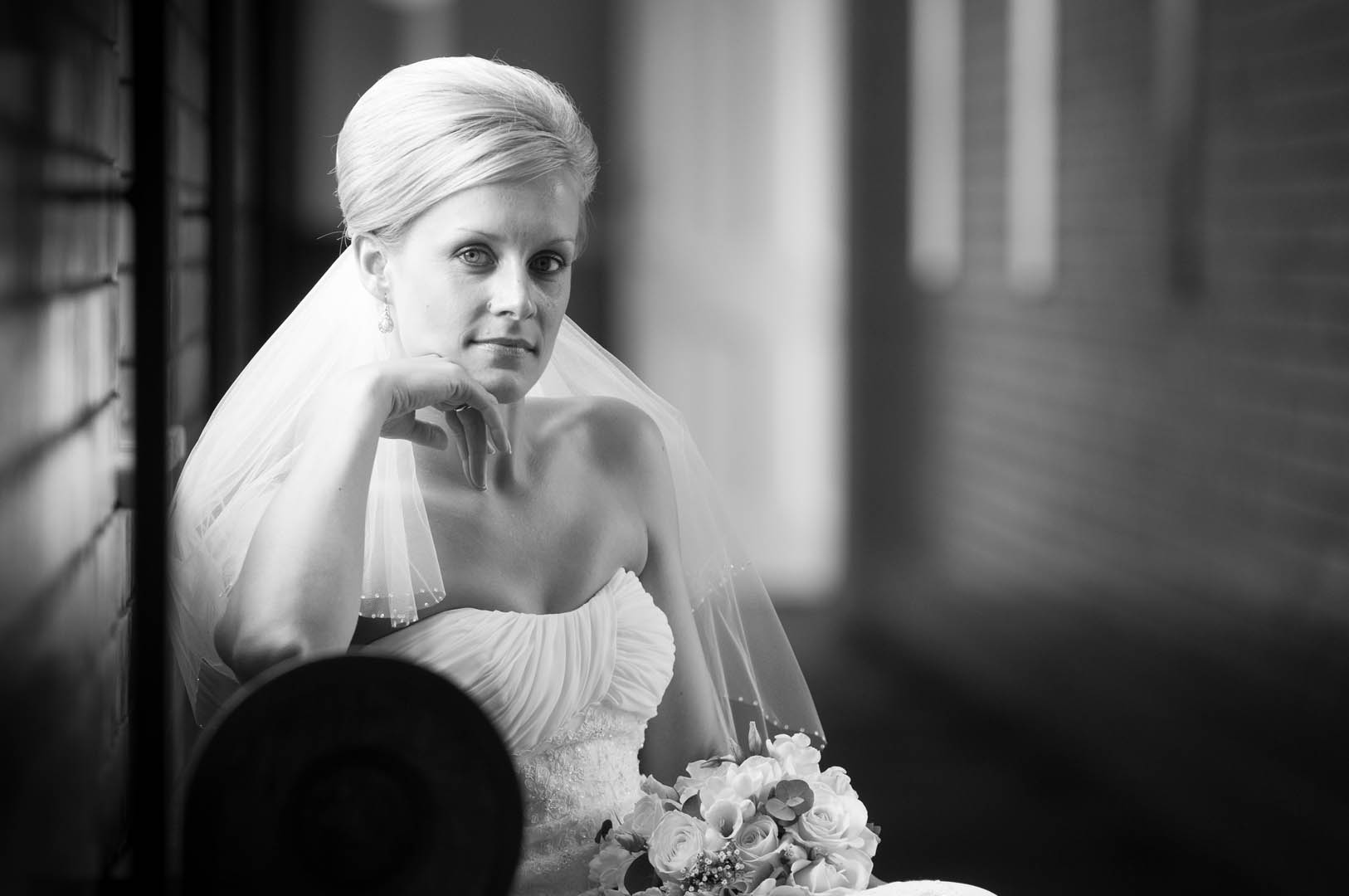 Bride in strapless gown, veil, holds flowers. Leans on hand, looks at camera. Black and white photo.