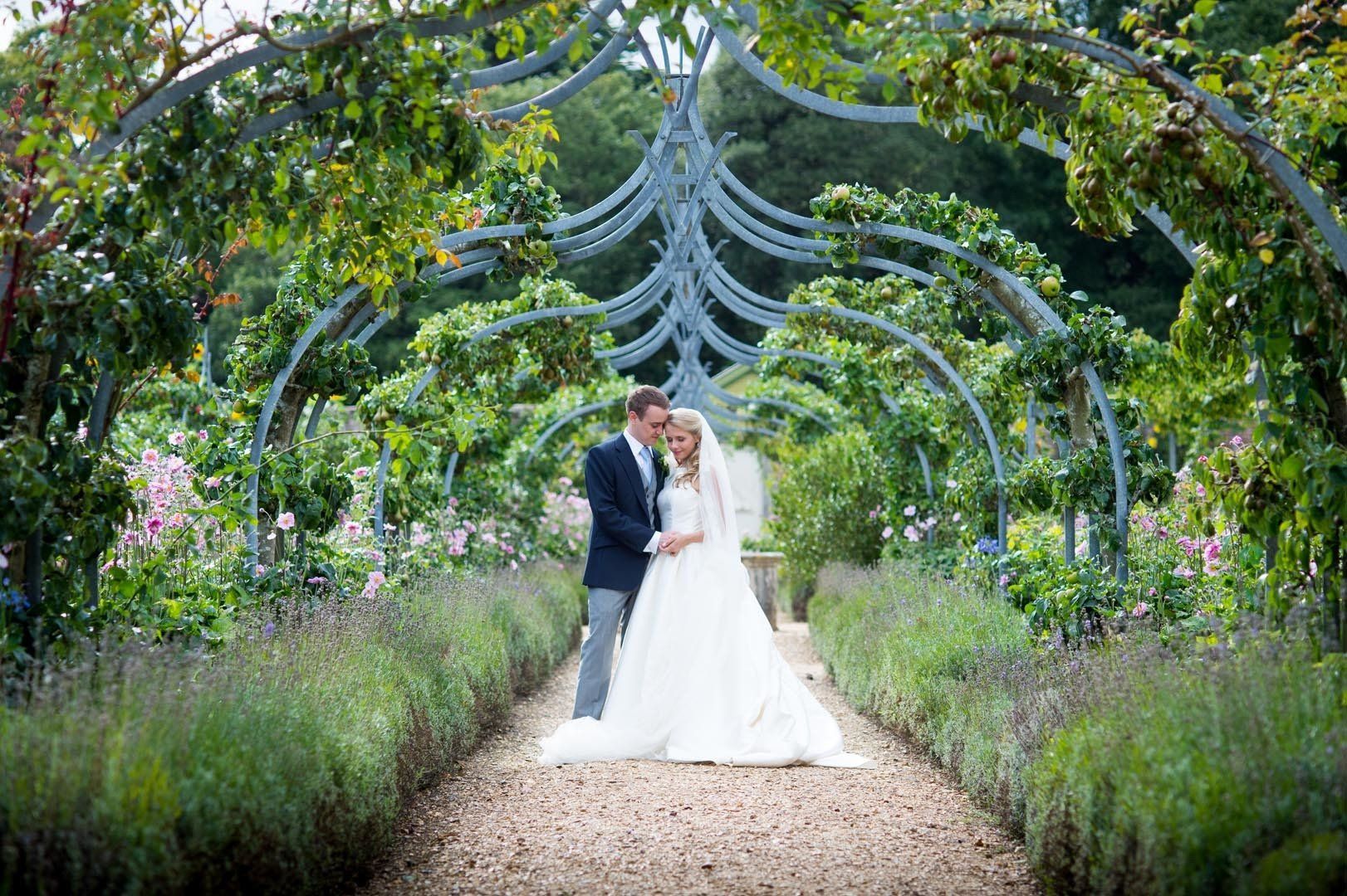 Couple embraces under a metal archway in a garden, bride in white gown, groom in suit.