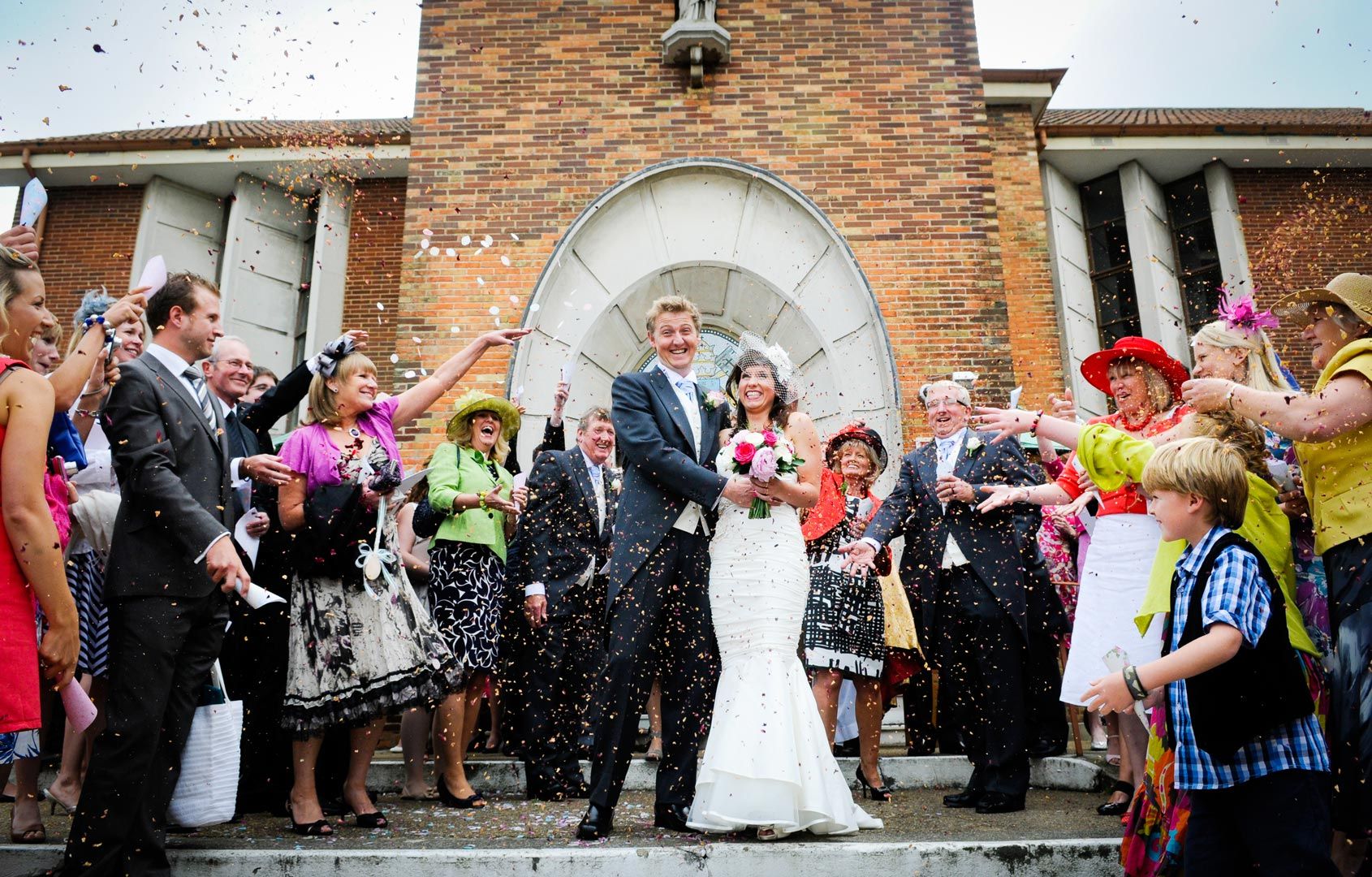 Bride and groom covered in confetti after exiting building, surrounded by wedding guests.