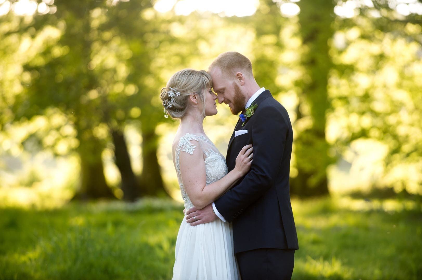 Bride and groom embrace outdoors, heads touching, surrounded by trees and sunlight.
