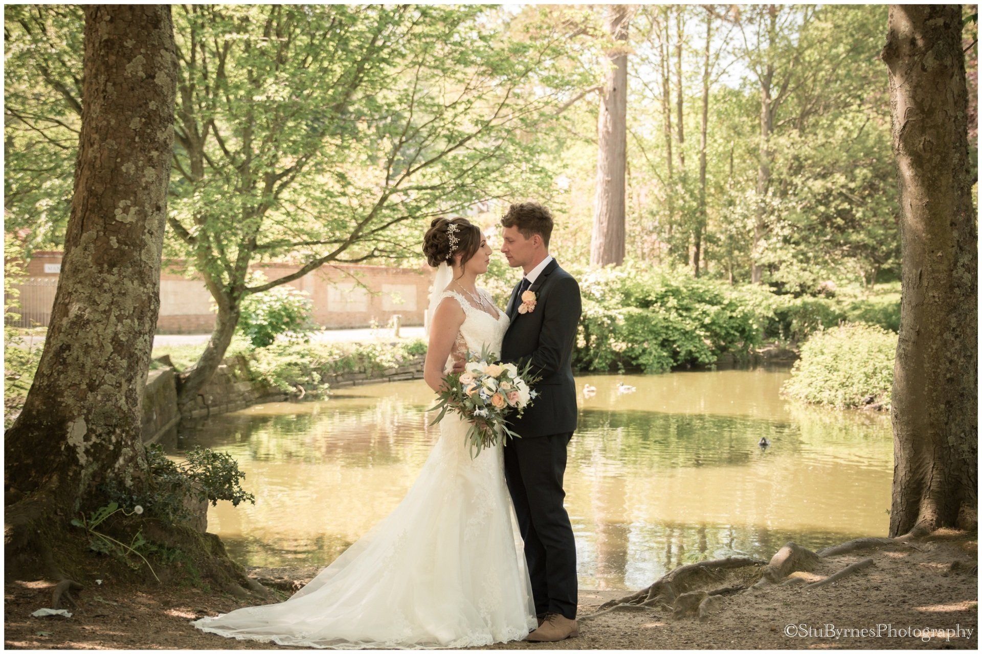 Bride and groom embrace by a pond in a park. She wears a white dress, he wears a suit. Sunlight filters through trees.