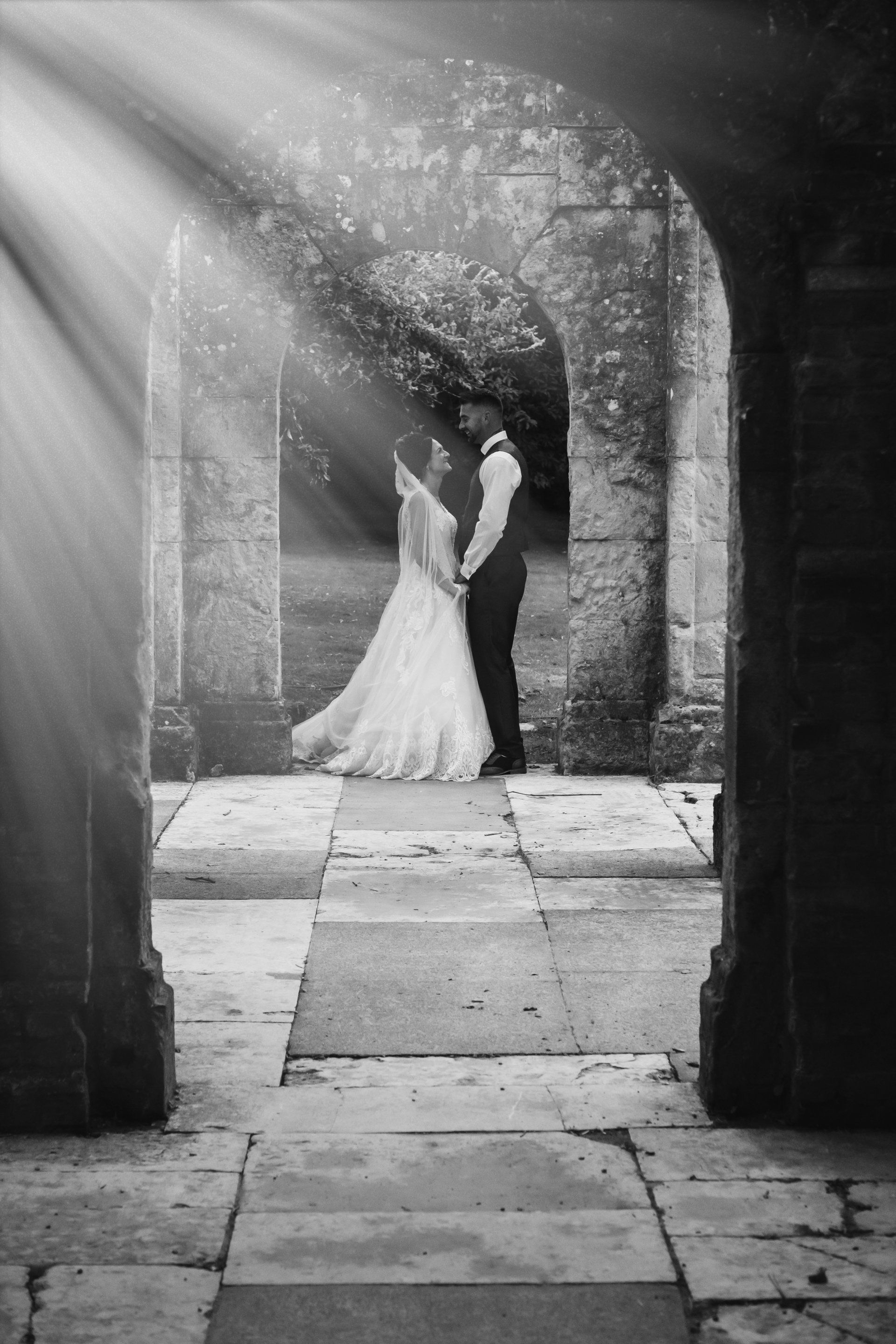 Bride and groom holding hands in stone archway, sunlight shining through.