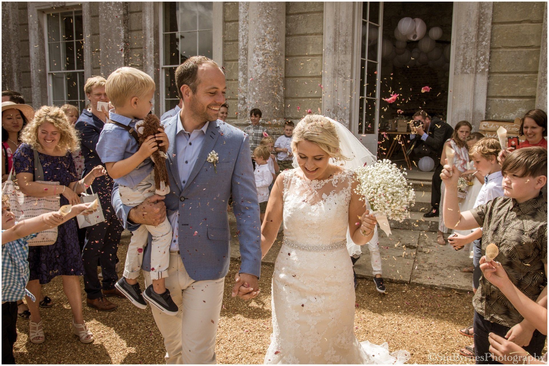 Newlyweds and child walk amid confetti, exiting a building. Light clothing, smiles, and celebrations.