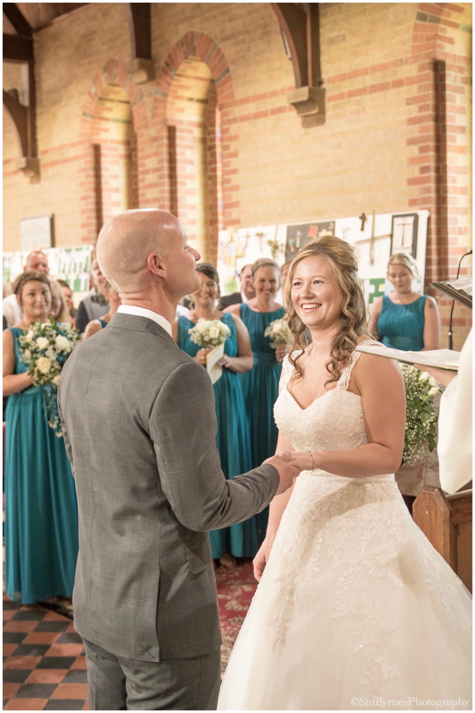Groom holding bride's hand at altar, exchanging vows. Bridesmaids in teal dresses watch in a church.