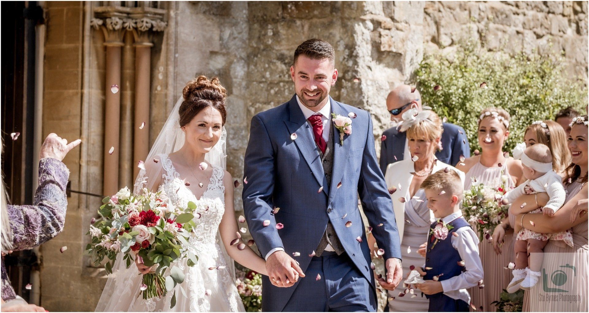 Newly married couple exiting a stone building, showered with confetti. Bride in white dress, groom in blue suit.