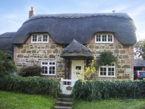 Stone cottage with thatched roof, white-framed windows, and a small porch entrance, green hedge in front.