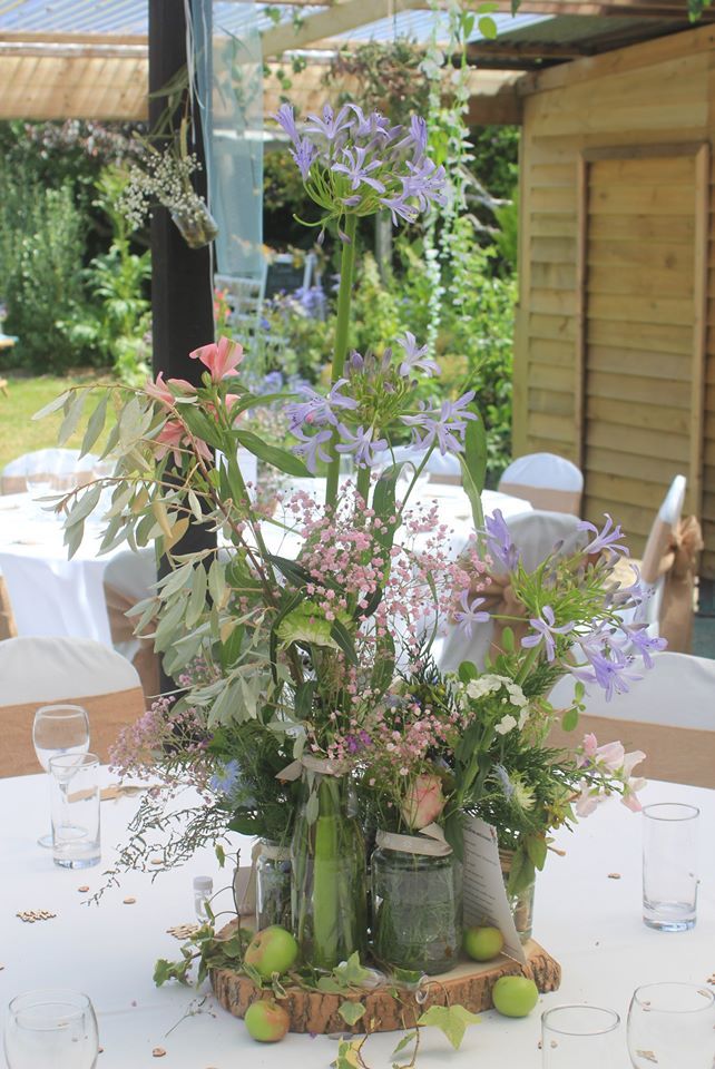 Floral centerpiece on a table, featuring blue and pink flowers in glass jars on a wood slice.
