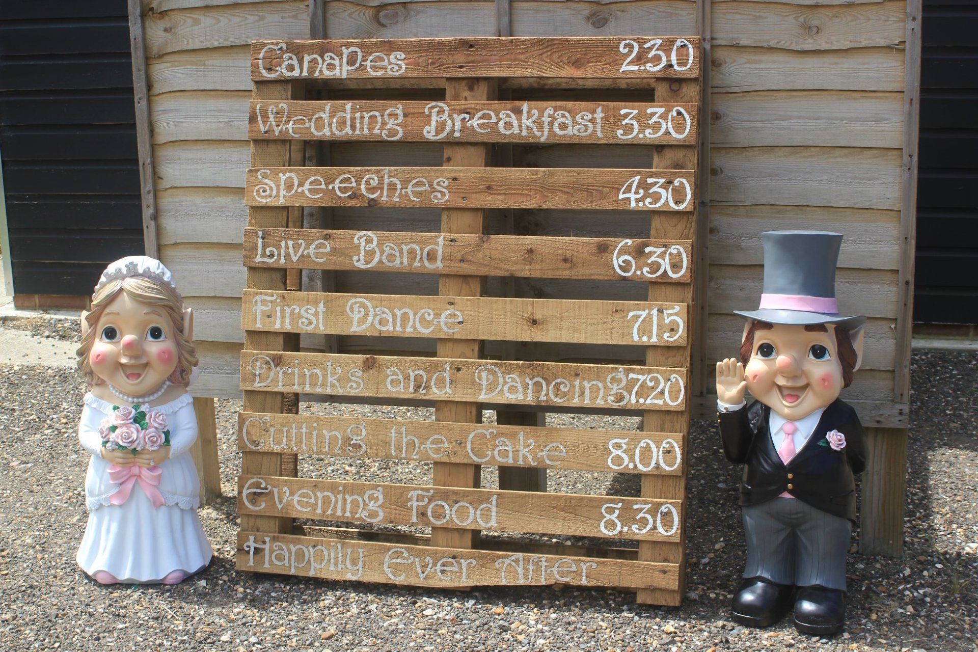 Wedding schedule on a wooden pallet with bride and groom statues.