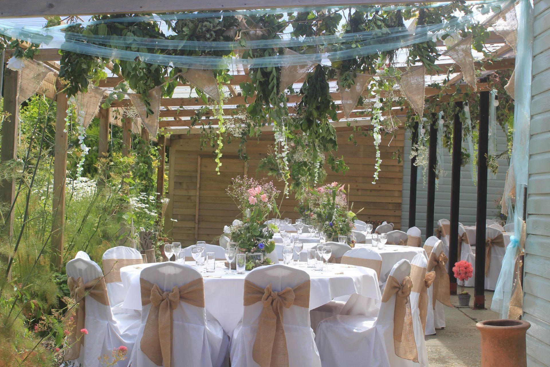 Outdoor wedding reception table, set under a pergola, with white tablecloths and rustic chair decorations.