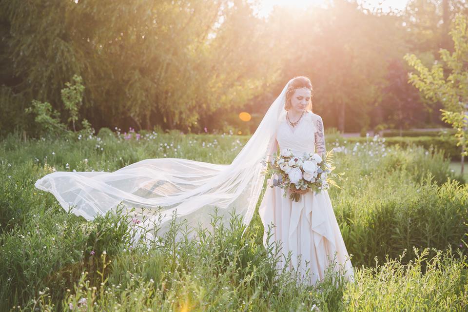 Bride in a flowing white dress and veil, holding a bouquet in a field of tall grass with the sun shining.