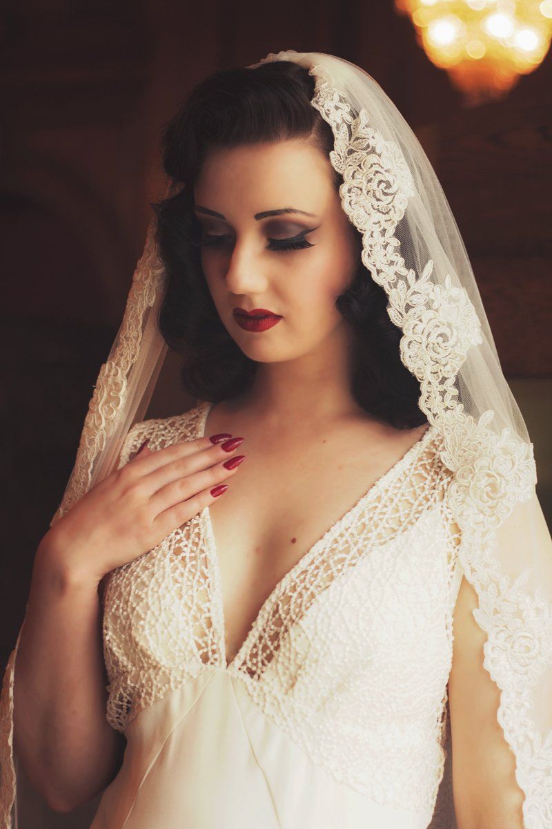 Bride in white lace wedding dress, veil; hand on chest, looking down, red lipstick.
